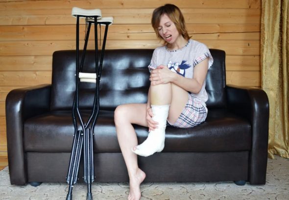 Full body of woman with aching leg sitting on couch near pair of crutches